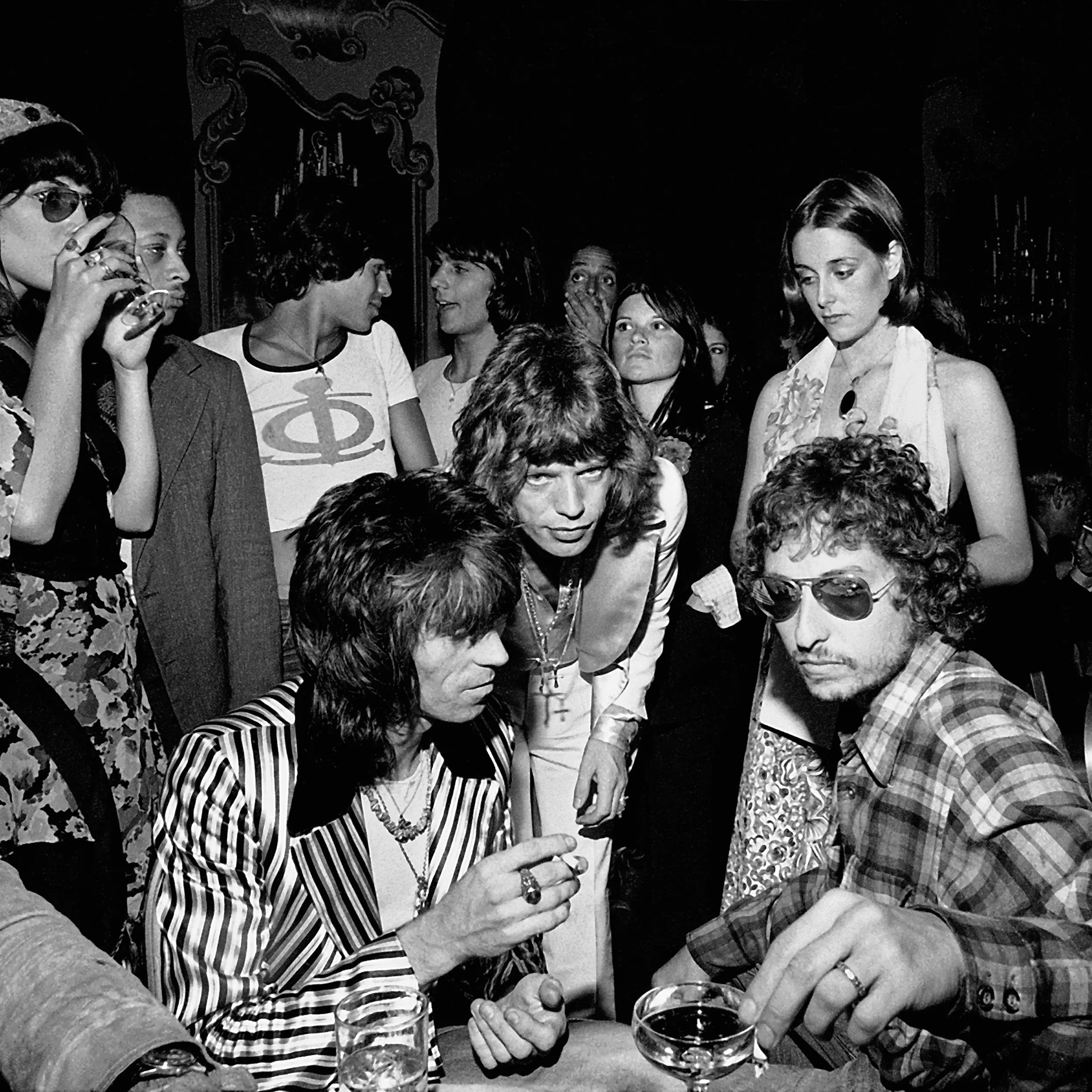 A black and white photo of a crowded party scene, with men in bold outfits and sunglasses in the foreground, surrounded by women and other guests, some holding drinks and looking in different directions.